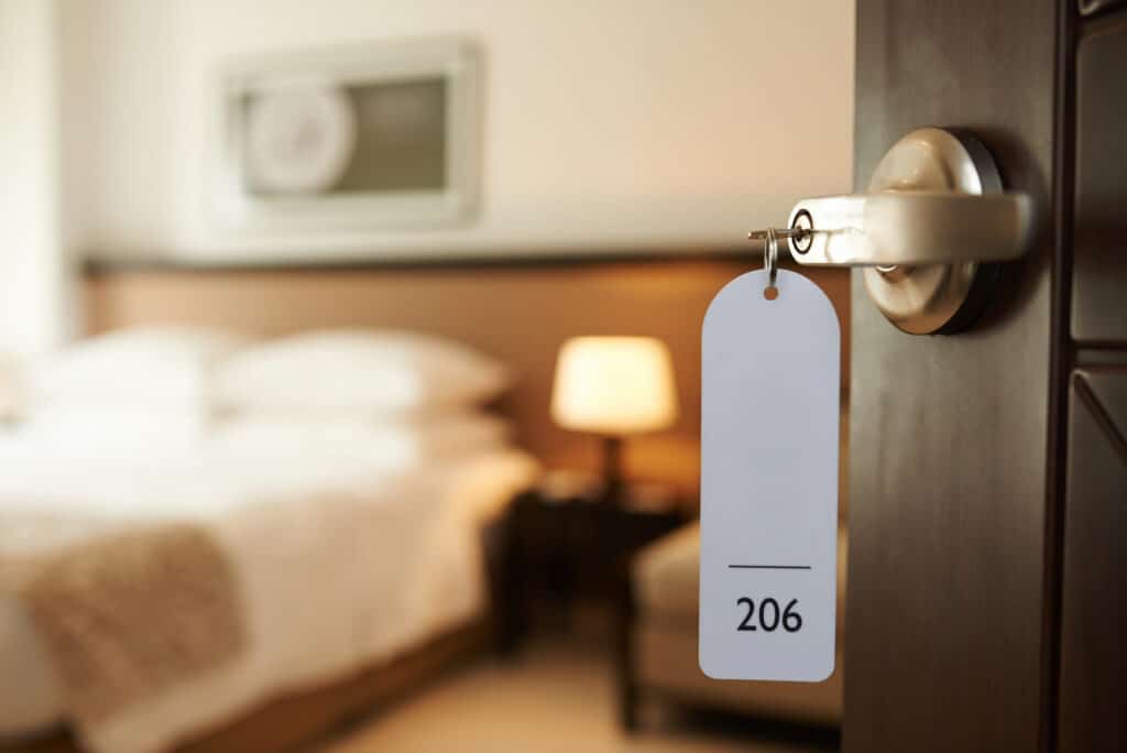 L'image montre une clé insérée dans la serrure d'une porte de chambre d'hôtel, avec un porte-clé numéroté 206 en gros plan. La chambre à l'arrière-plan est floue, mais on peut discerner un lit double avec des draps blancs et une lampe de chevet allumée, ce qui crée une atmosphère accueillante et confortable. L'éclairage doux et la décoration intérieure suggèrent un environnement de repos et de détente.
