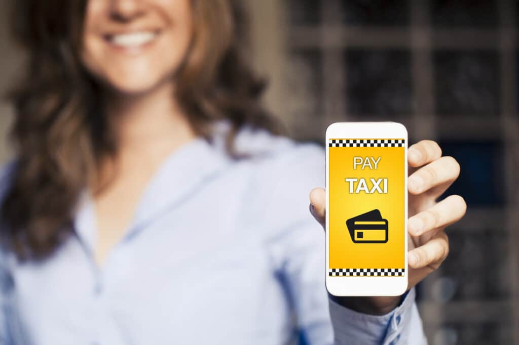 L'image montre une femme souriante tenant un smartphone avec un écran affichant une application de paiement de taxi. L'application semble avoir une interface jaune avec un motif de damier de taxi et une icône de carte de crédit, suggérant une fonctionnalité pour payer un service de taxi directement depuis le téléphone. La mise au point est sur l'écran du téléphone, rendant le visage de la femme et l'arrière-plan intentionnellement flous pour attirer l'attention sur l'application.